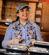 Zoey Al-Dahash in a denim cap and floral-embroidered pinstripe shirt smiling at an unseen customer from behind a counter covered with to-go containers